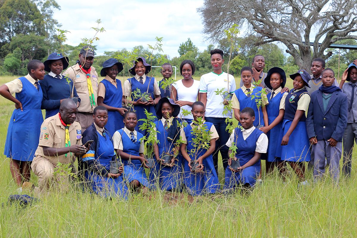 A group of children in blue school uniforms, with some adults, in a field with trees behind. Some are holding saplings.