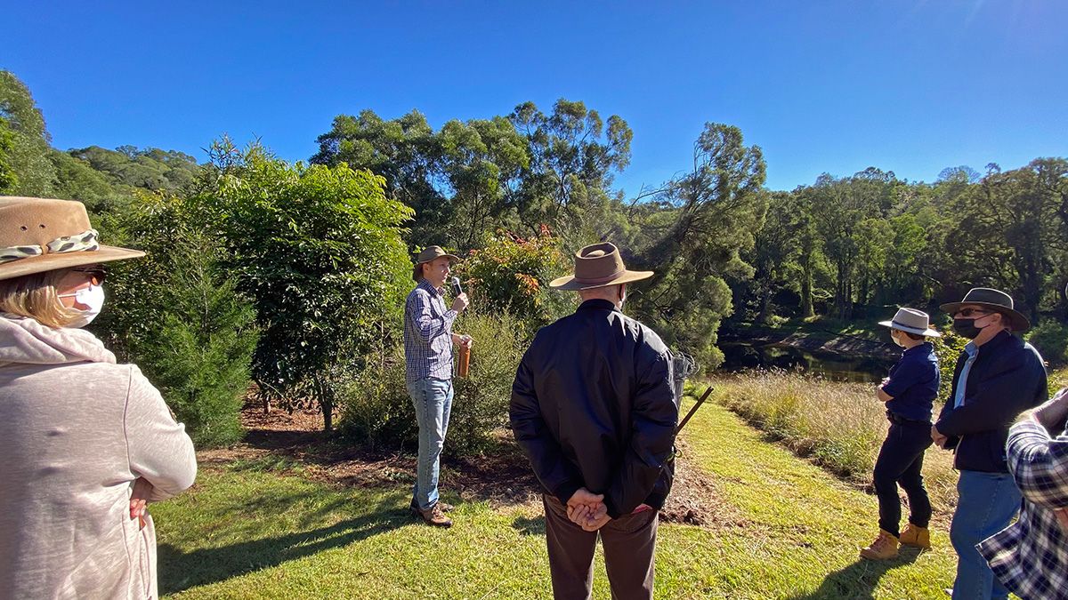 Five individuals wearing masks stand around a man with a microphone giving a talk, outdoors on a grassy area surrounded by trees.