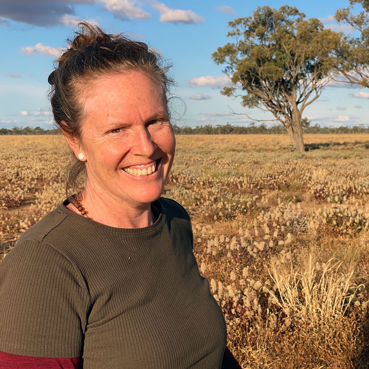 Deborah Metters smiling in front of a landscape of golden scrubby grasses with a tree in the background.