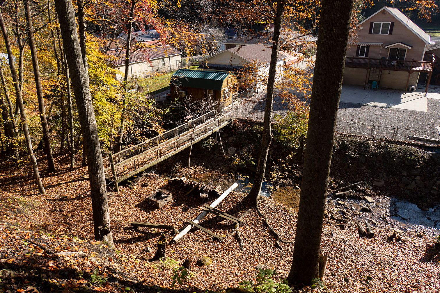 A footbridge across a creek on a sunny day. On one side are houses; on the other, trees.