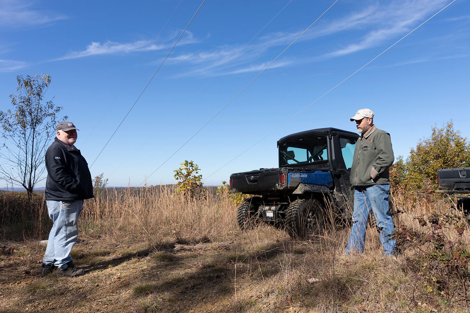 Two men stand on either side of the frame in a brown grassy area with blue sky behind.