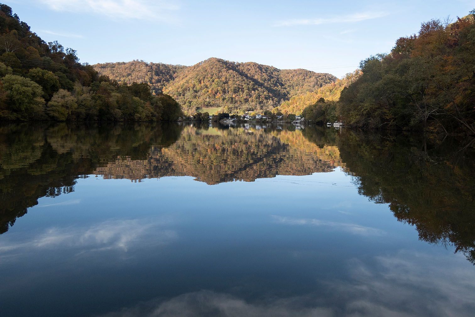 Tree-covered hills and a cloud-scattered sky reflected in calm water.