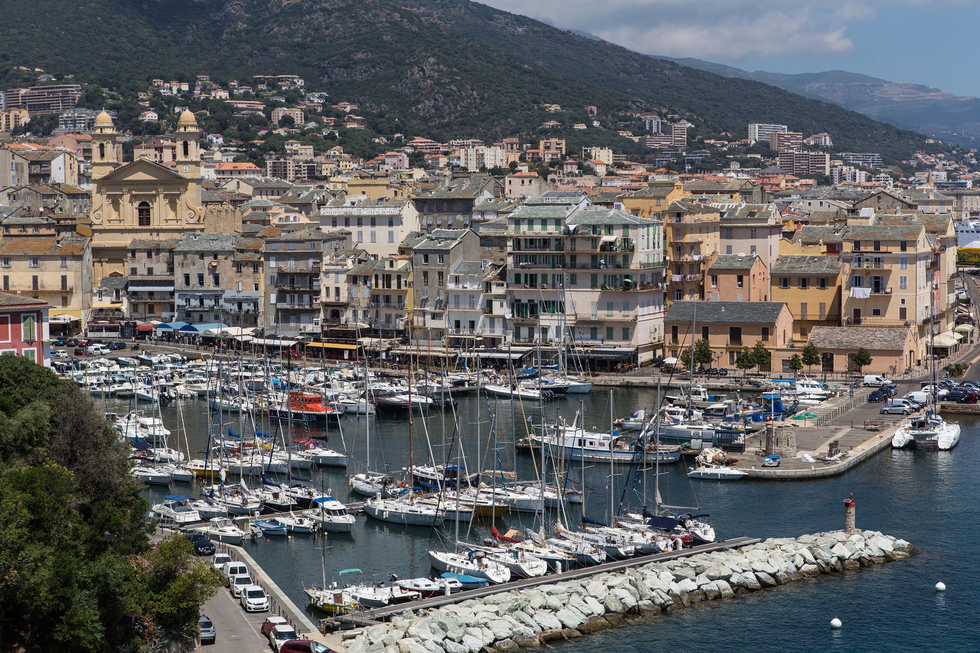 The harbour in Bastia, Corsica. Boats are moored and buildings and hills visible behind them.