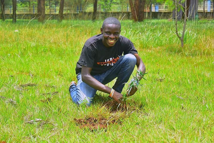 Kenyan student Lamech Opiyo crouched in the grass holding a newly planted seedling