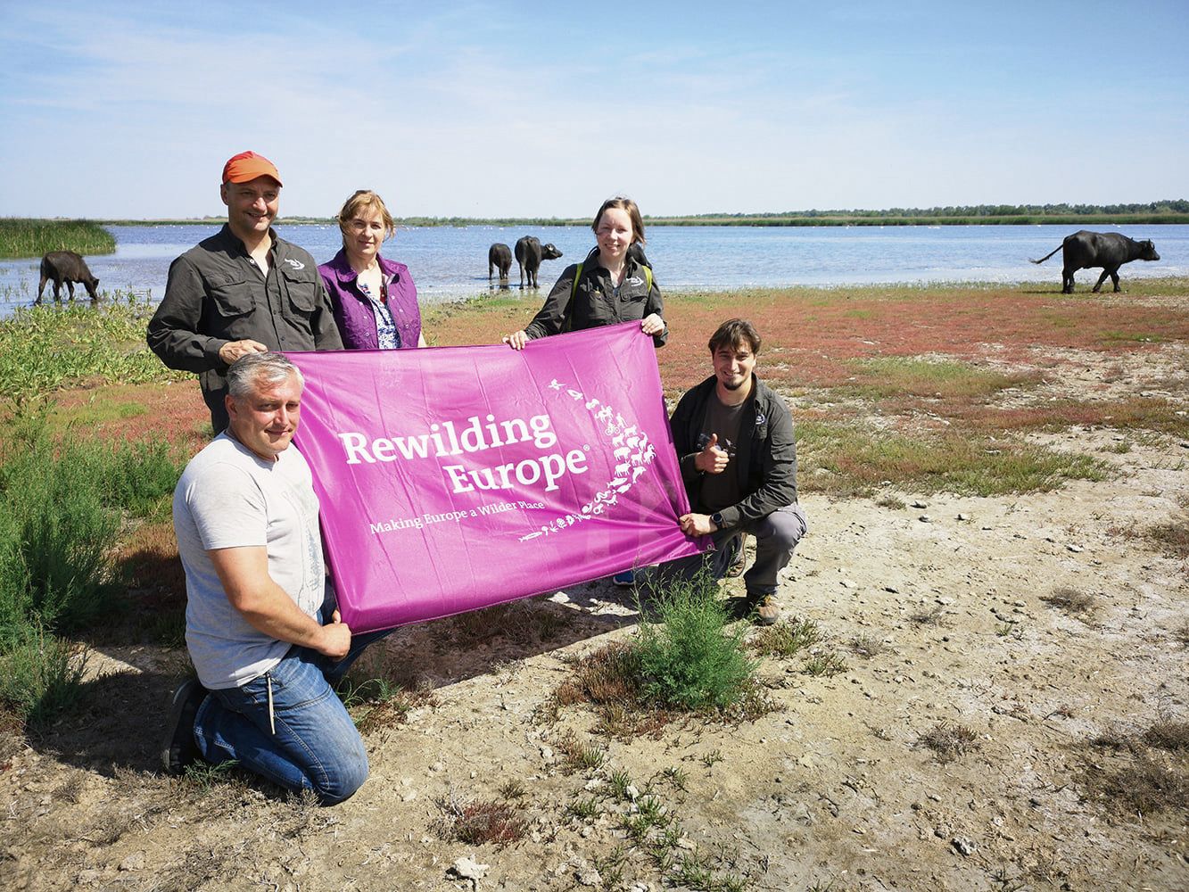 Five people hold a banner that says "Rewilding Europe" in a river landscape with animals