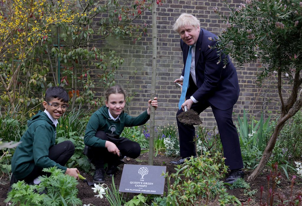 Two children, crouched, smile for the camera in a garden, while Boris Johnson stands with a shovel full of dirt