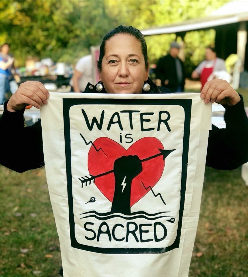 Christi Belcourt holding up a painting of a fist holding an arrow over a heart with the words "water is sacred"
