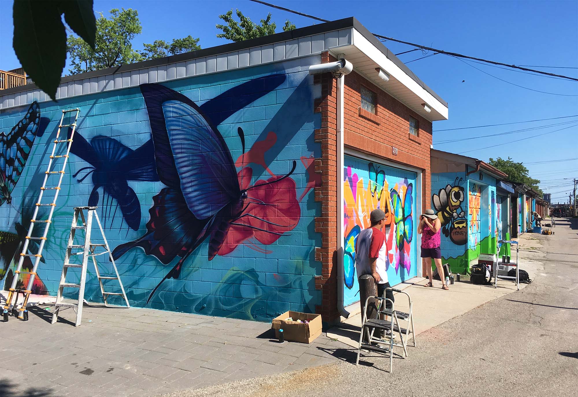 A series of garages in a laneway being painted with butterfly art on a sunny, blue-sky day 