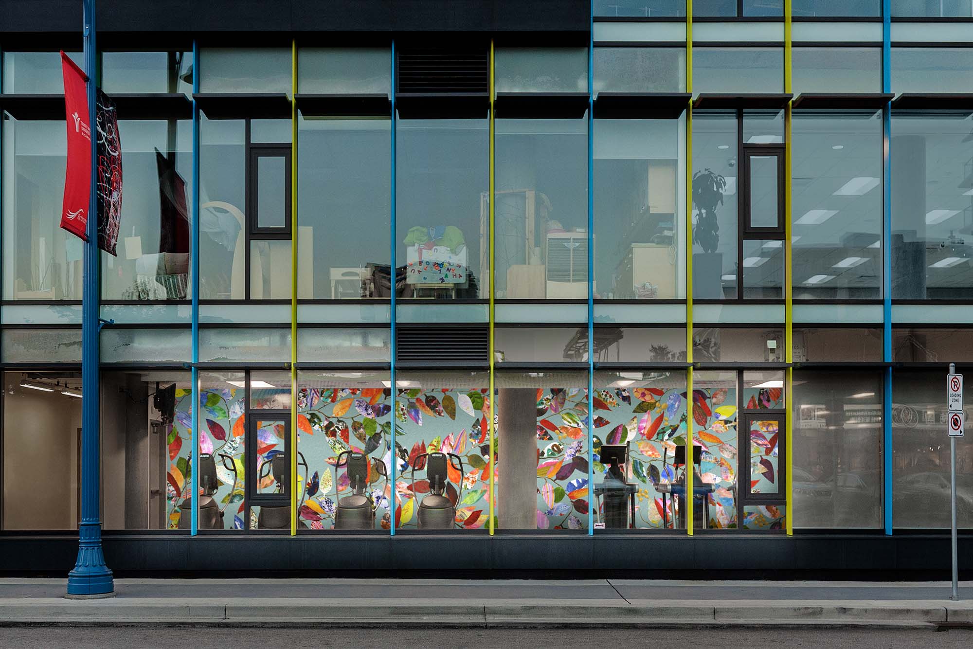 View of the exterior of a glass-walled building. On the ground floor, a mural of leaves is visible behind fitness machines.