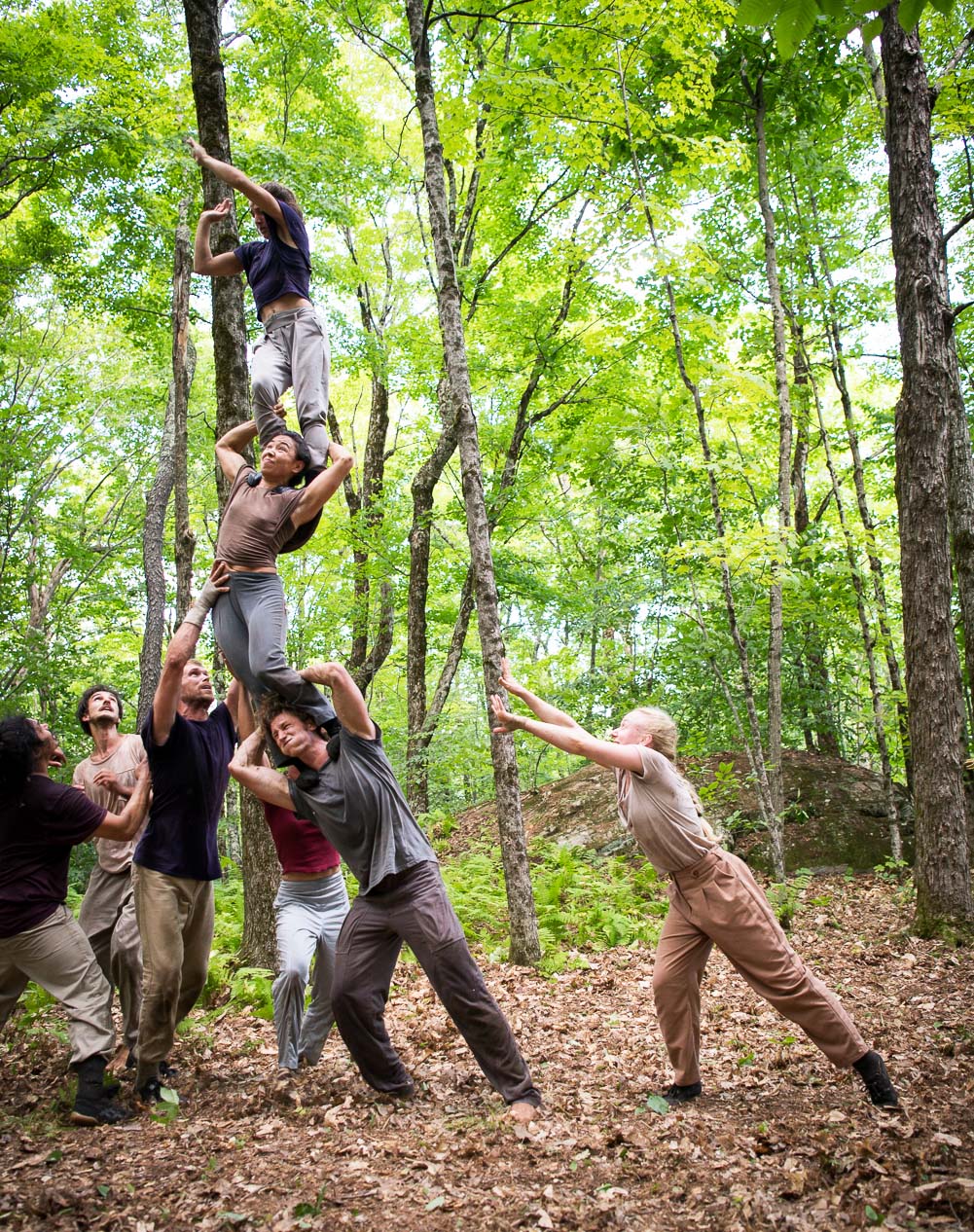 In a forest, one group of performers holds a person high overhead while another reaches to save them