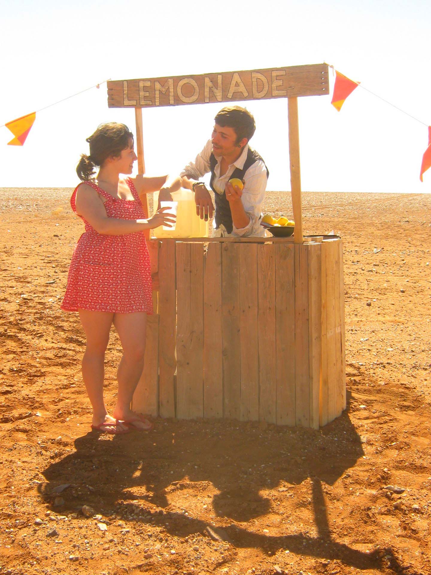 Two people at a wooden lemonade stand in a barren, reddish, rocky landscape