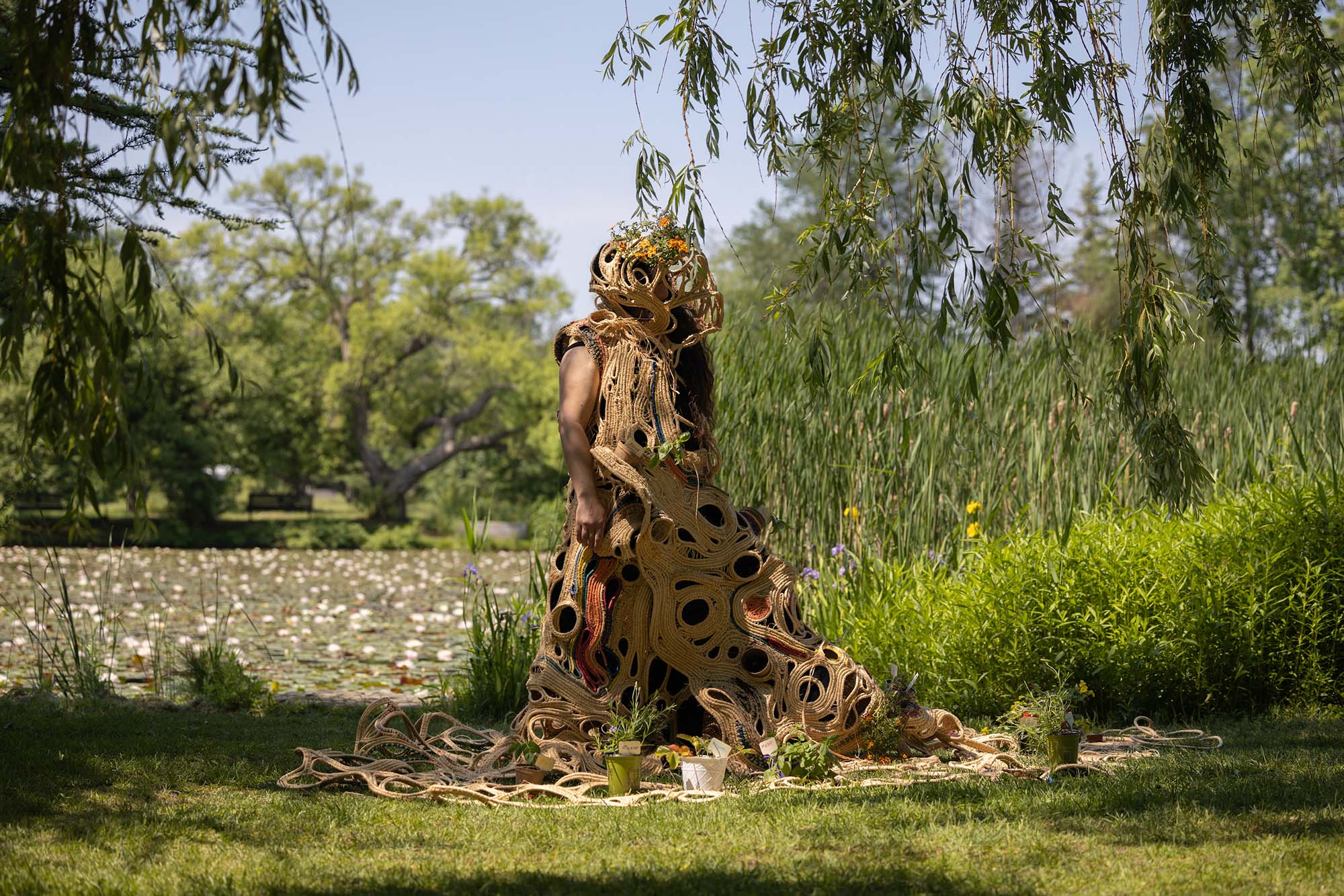 a performer draped in a sculpture of rope in the middle of a garden, surrounded by trees and plants