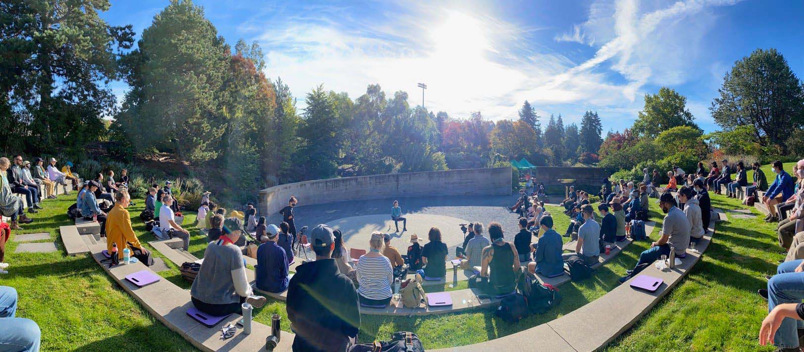On a sunny day, a large group of people sit around an open-air amphitheatre