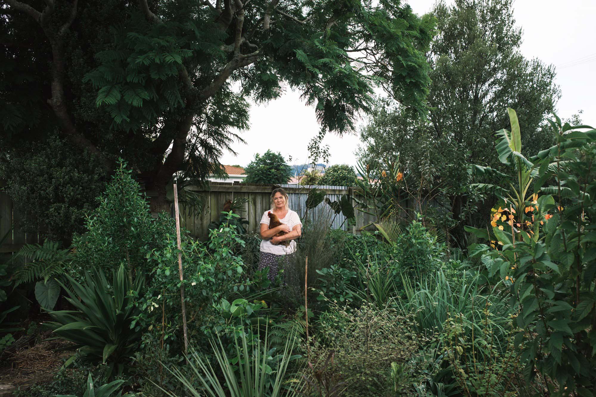 A person holding a chicken, standing in a lush green garden