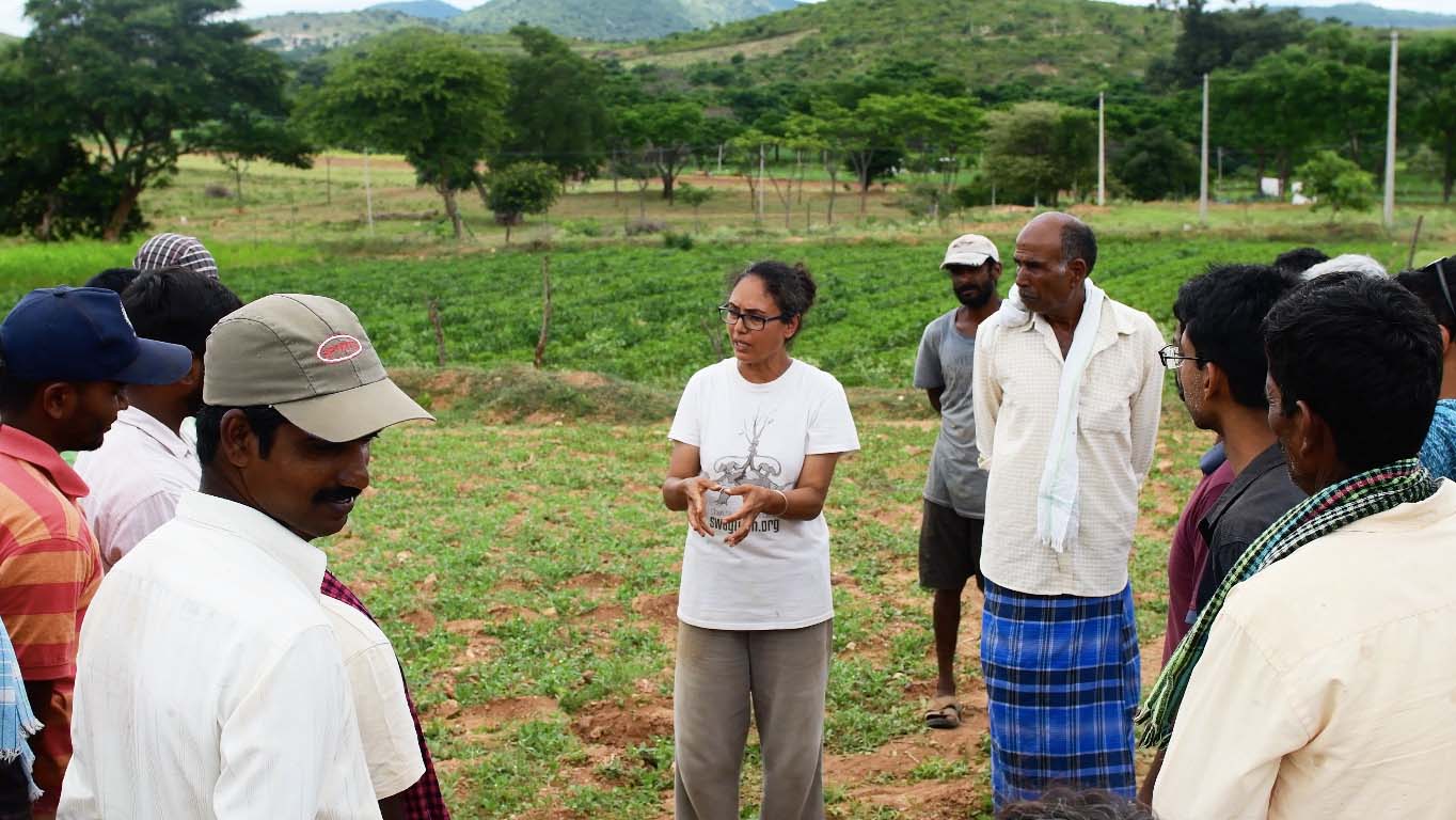A person talking to a group of people while standing outdoors, with crops and trees in the background