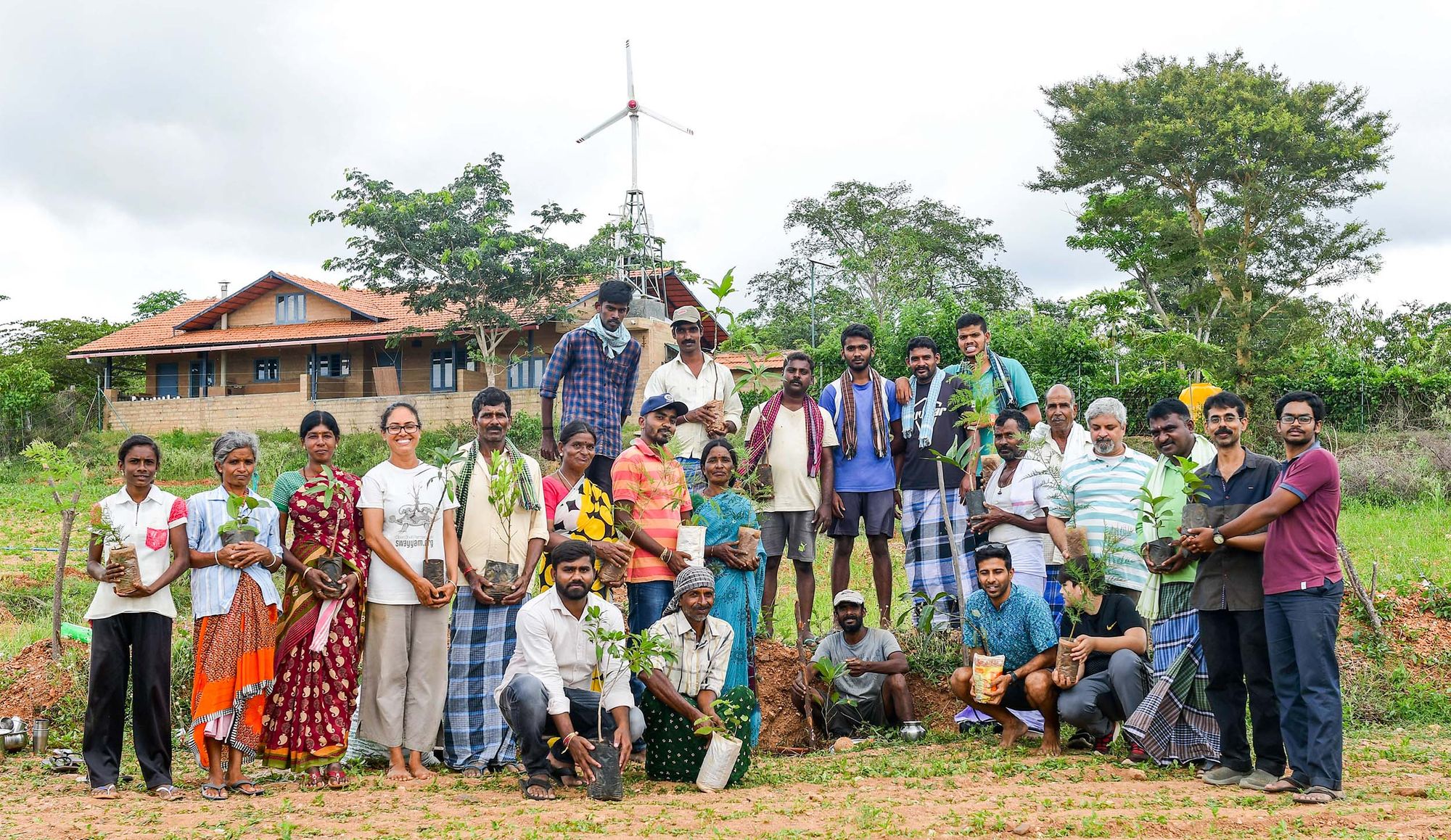 A group of people holding potted tree seedlings, posing outdoors in front of a house and trees