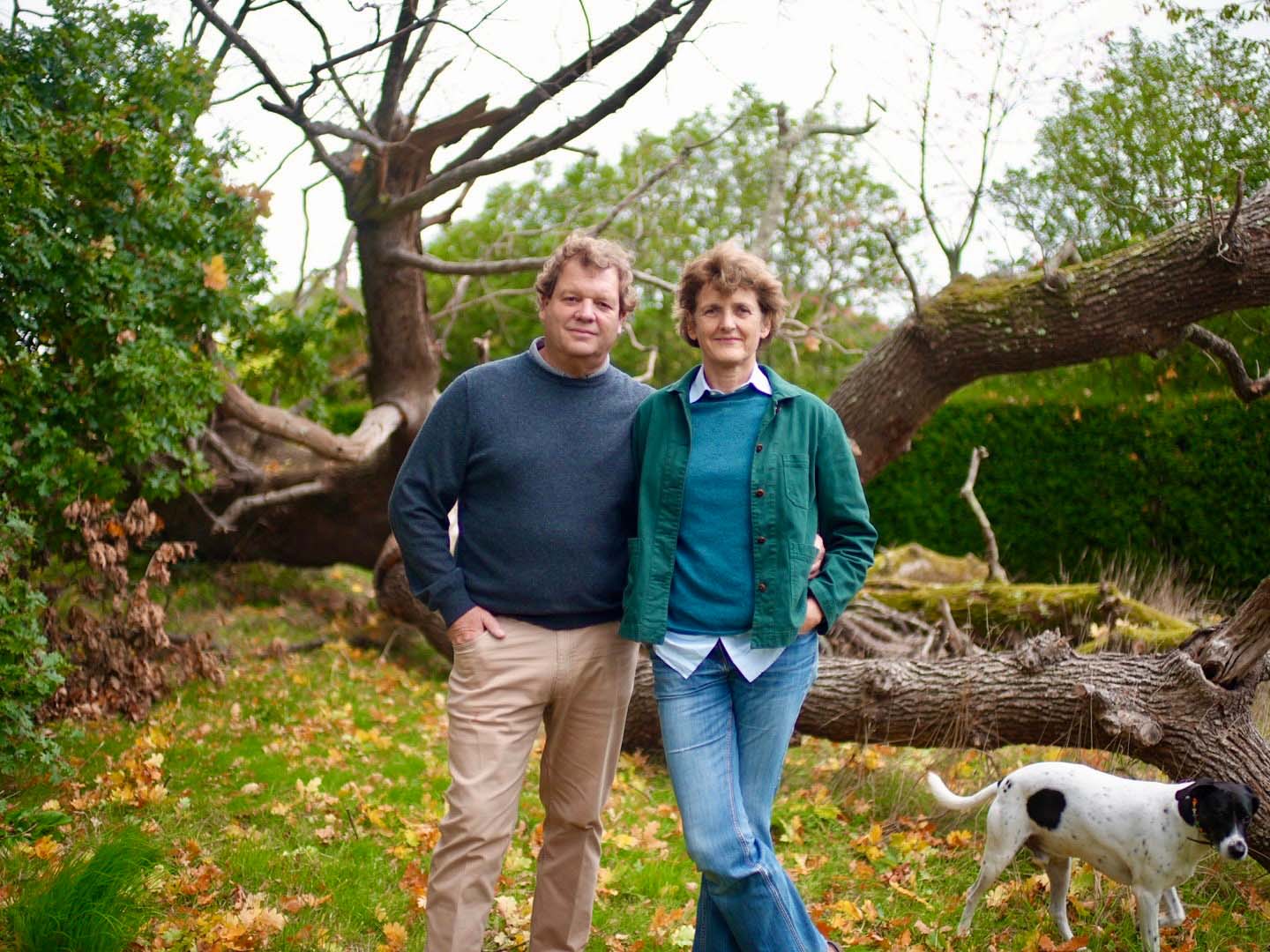 Two people standing outdoors in front of a fallen tree. A dog is walking around next to them
