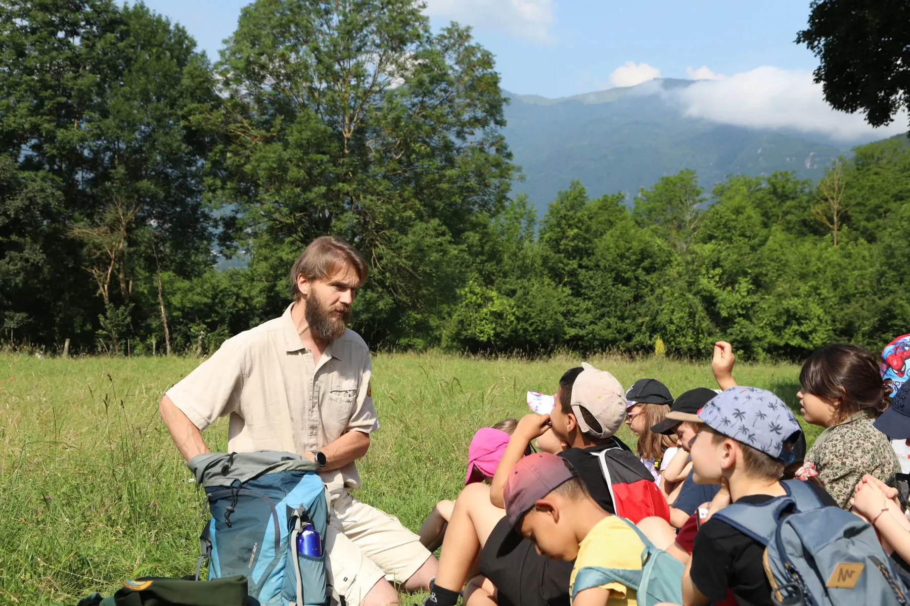 In a field near a forest, a person holding a backpack speakers to a group of children