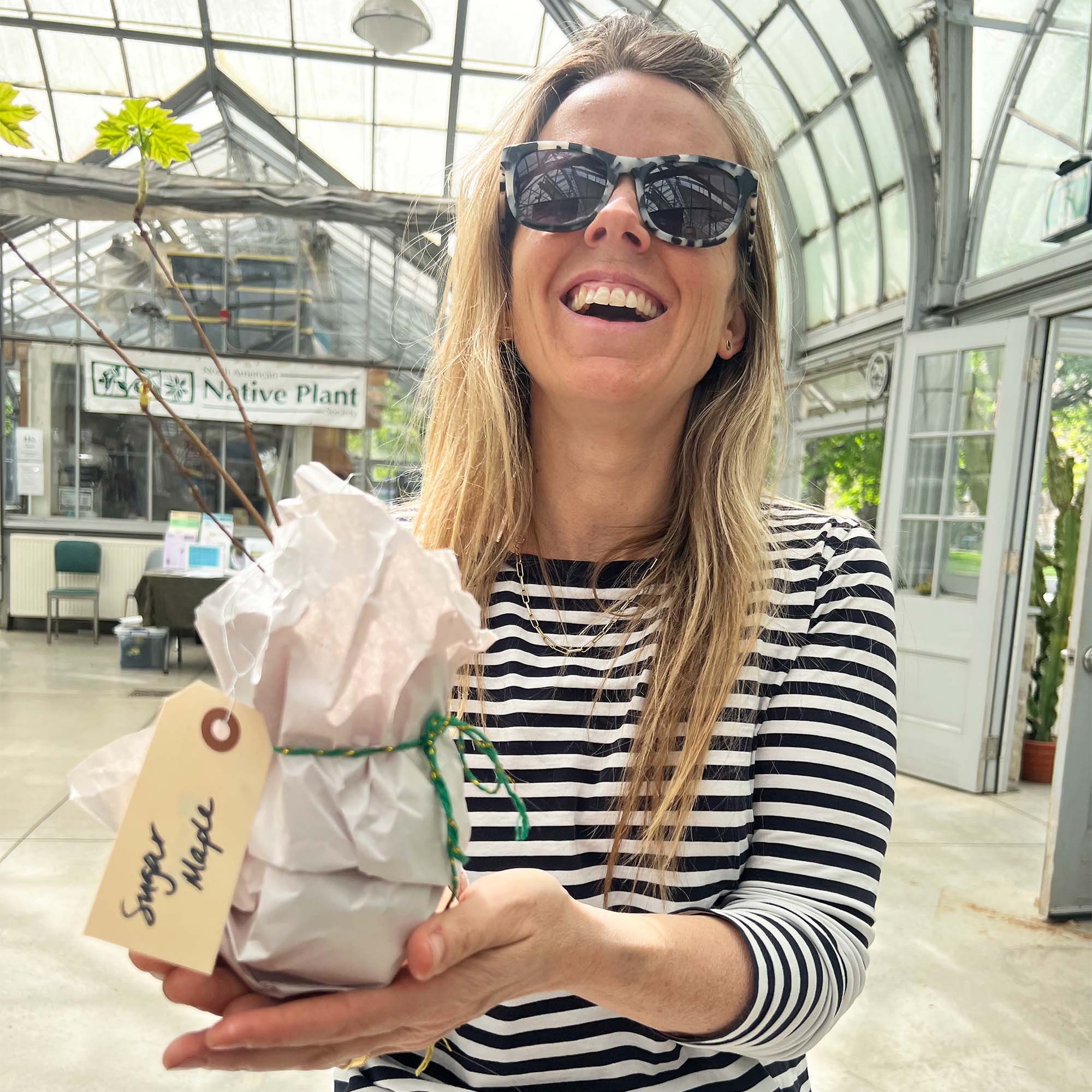 A smiling person in sunglasses holding a tree seedling at a nursery