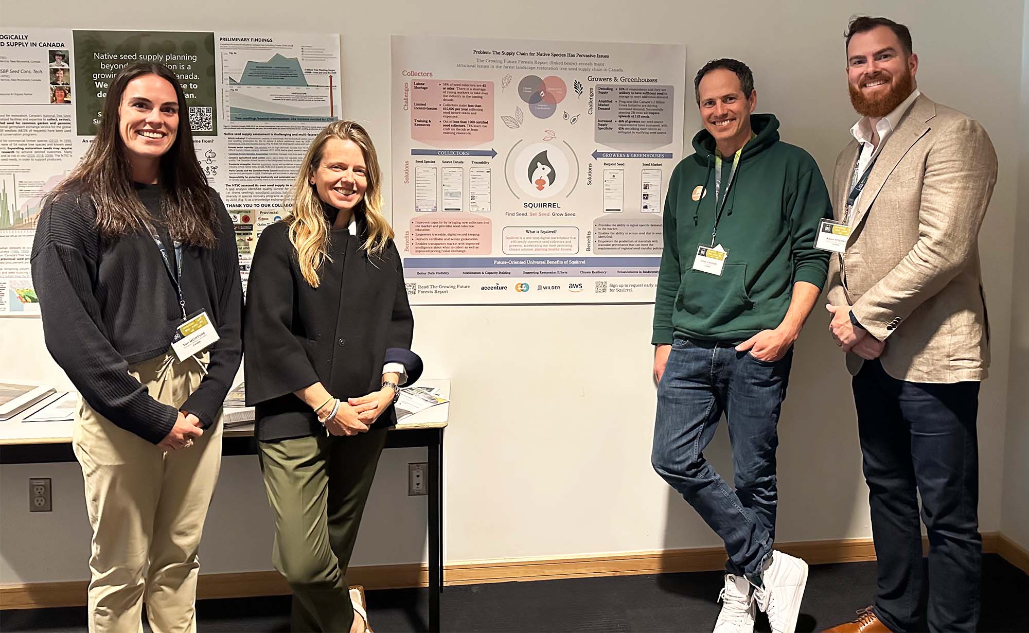 Four people wearing conference lanyards posing and smiling for the camera in front of wall-mounted posters