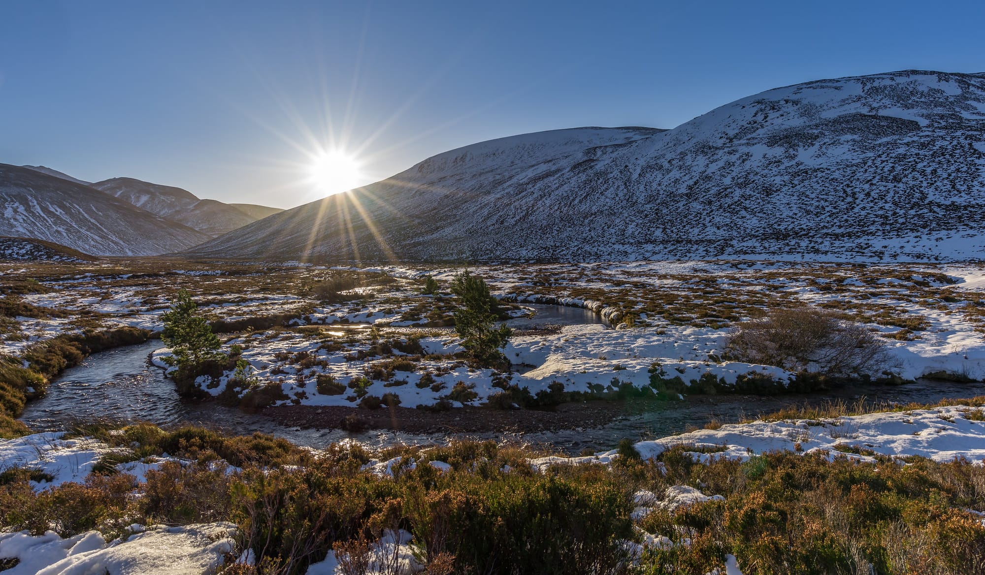 A river curving through a hilly landscape with snow, and the sun just over the horizon in a blue sky