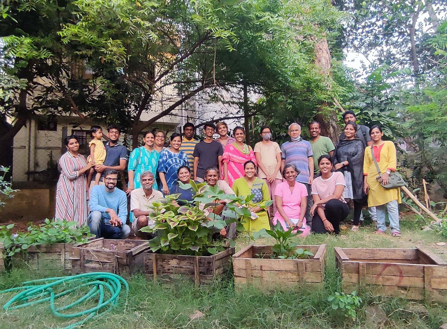 A group of people posing under a tree with wooden planter boxes and grass in front