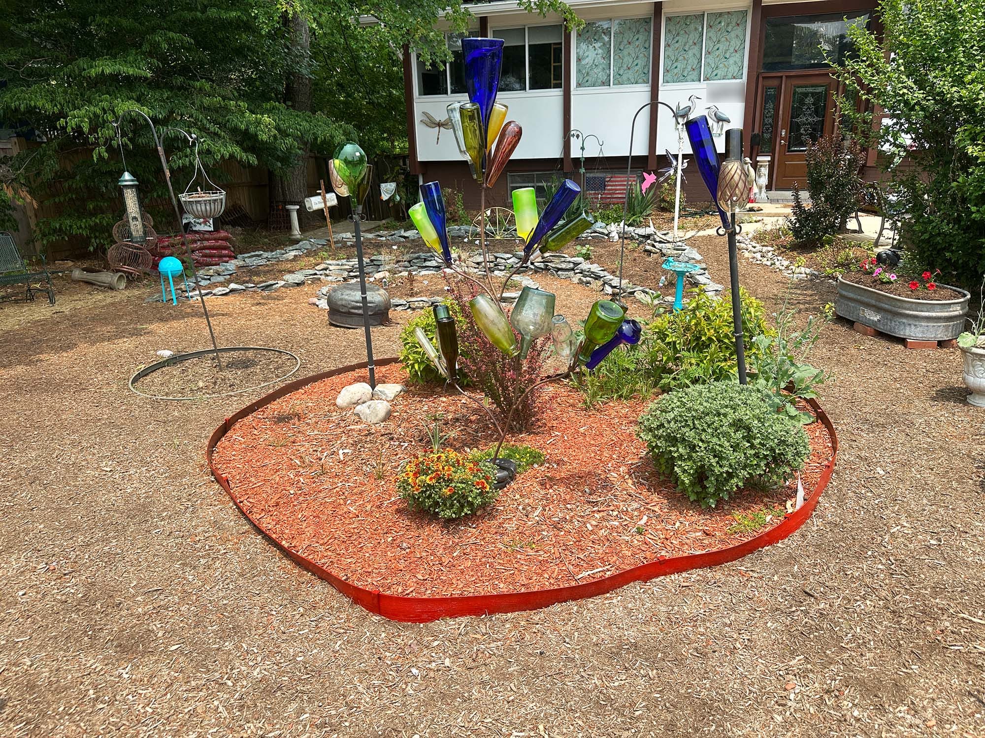 A garden bed outlined in red, filled with plants and a bottle sculpture and surrounded by wood-chip mulch