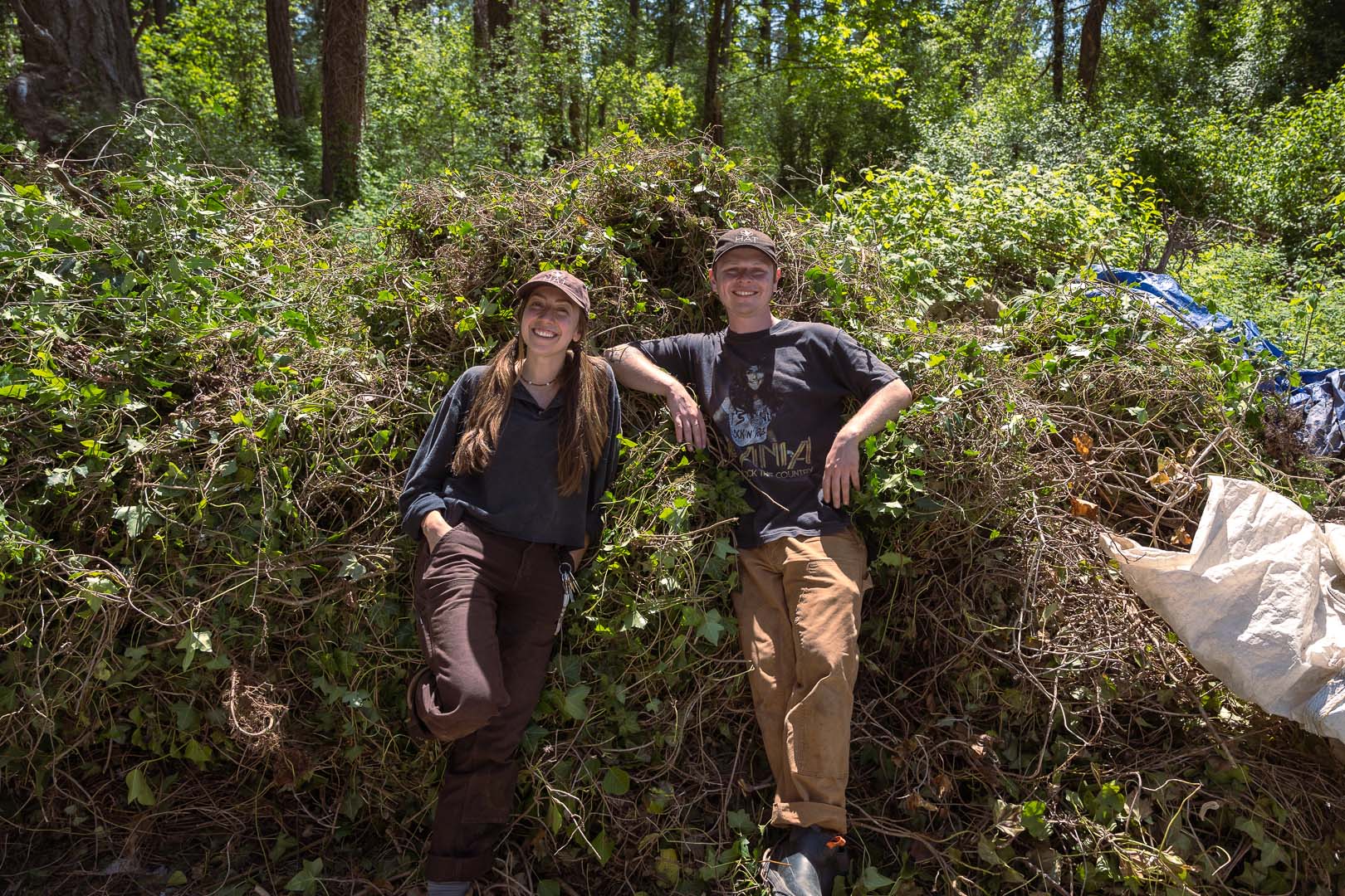 Two people smiling, leaning against a dense thicket of greenery
