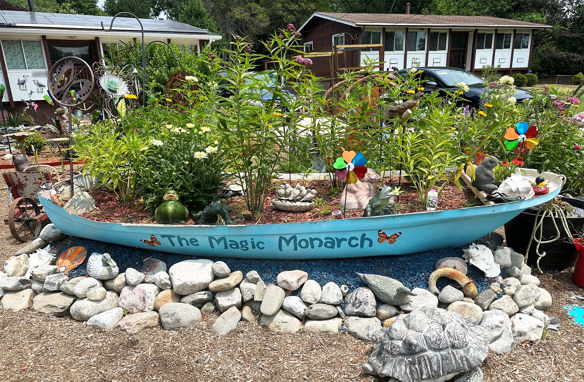 in the front yard of a house, a blue-painted boat with the words "the magic monarch" is being used as a planter