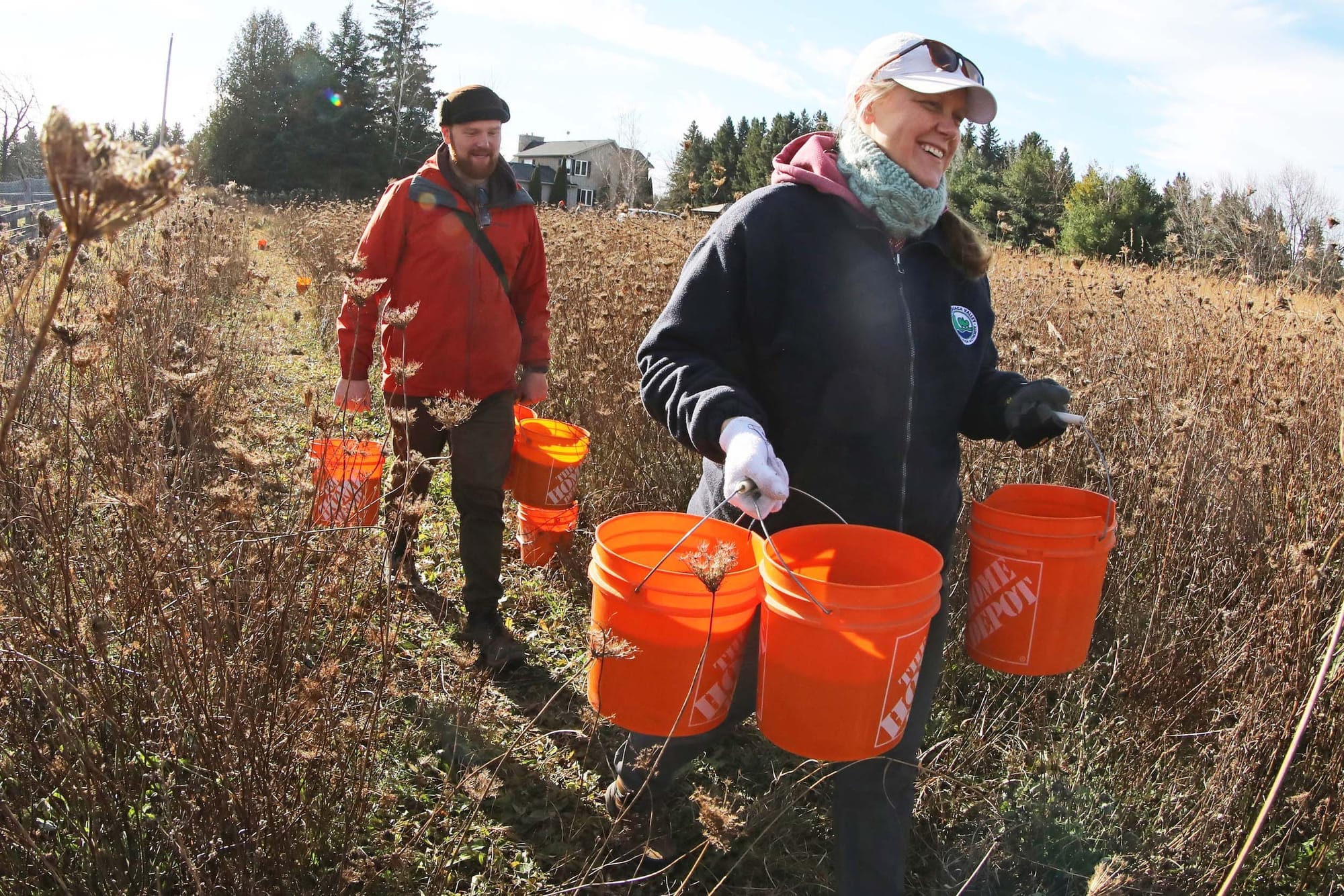 Two people carrying buckets in a field of dried grass and plants