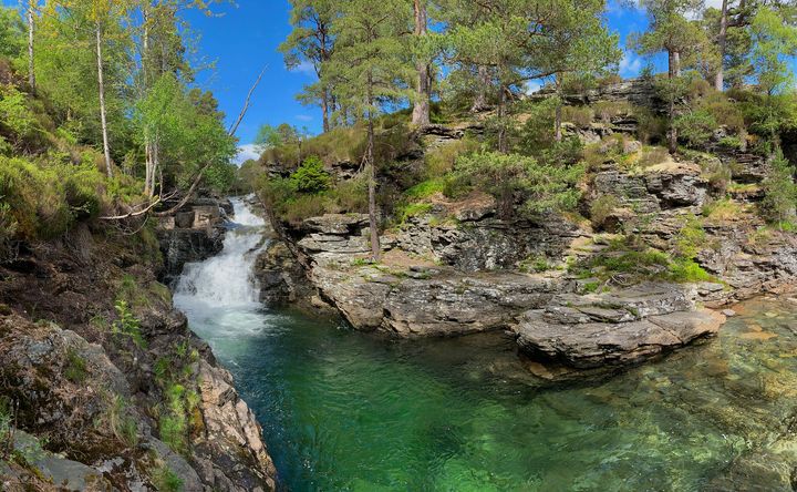 A waterfall and blue-green river run past tree-covered rocky land