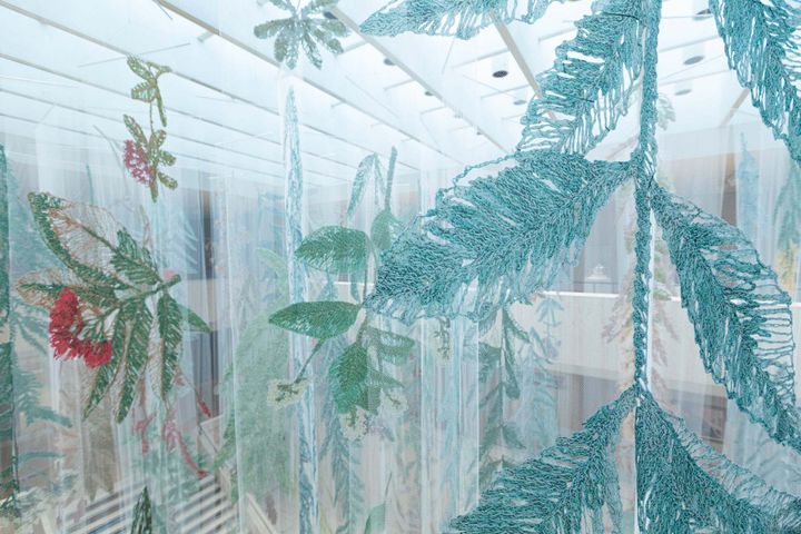 Large sketches of plants mounted on transparent fabric hang from the ceiling of a bright gallery