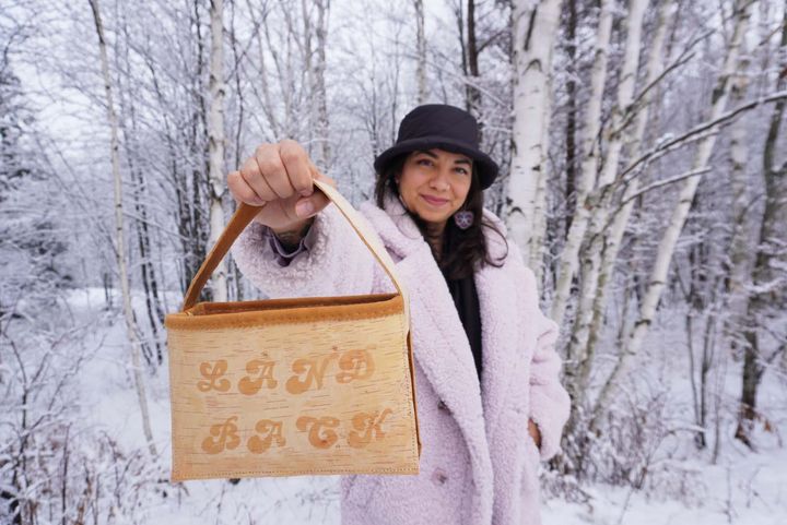 A person stands amidst birch trees in winter, holding out a birchbark bag that reads "Land Back"