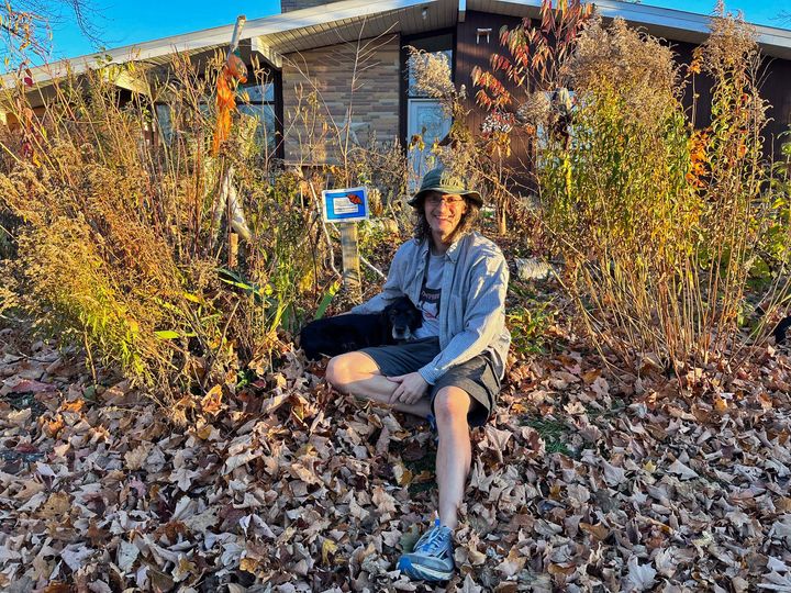 A smiling person sitting on the ground amidst dry fallen leaves and a garden, in front of a house