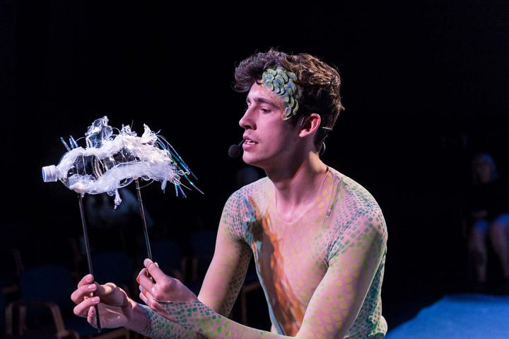 An actor on a stage wearing mermaid-like costumery and holding up a transformed plastic bottle