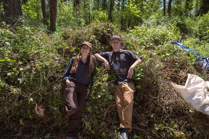 Two people smiling and posing for the camera, standing outdoors leaning on piles of English ivy